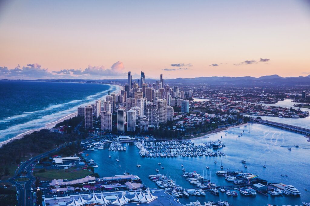 Aerial view of a coastal city with high-rise buildings, a marina filled with boats, and a long sandy beach beside a blue sea under a pastel sunset sky.