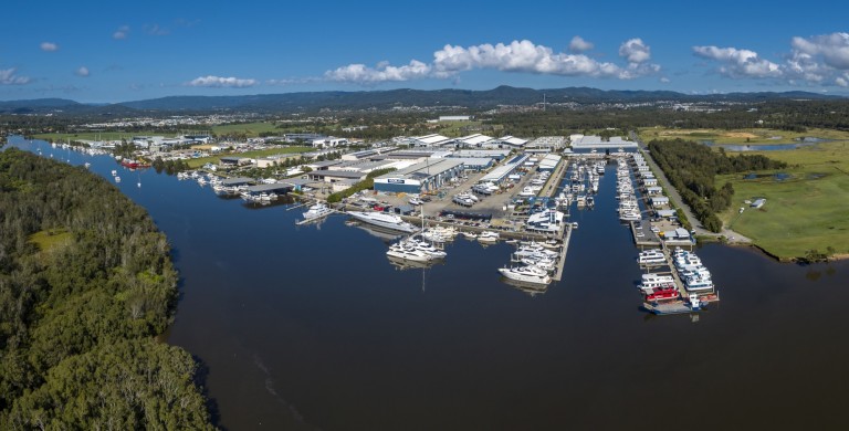 Aerial view of a marina with numerous boats moored along a river, surrounded by green trees, grasslands, and industrial buildings under a blue sky with scattered clouds.