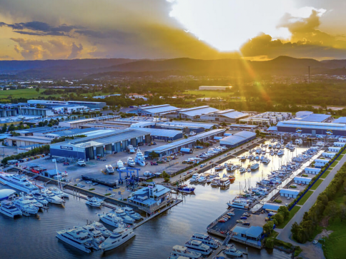 A marina at sunset with many moored boats and yachts, modern buildings, and warehouses nearby; sunlight streams over distant hills, casting a golden glow on the water and landscape.