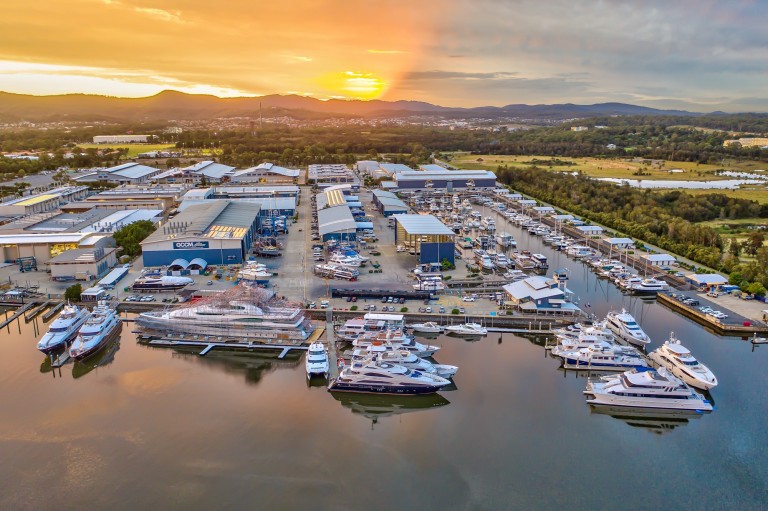 Aerial view of a marina at sunset, with numerous yachts and boats moored along the waterfront. Industrial buildings and green hills are visible in the background under a colourful sky.