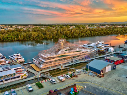 A large yacht wrapped in scaffolding is moored at a marina beside other boats, with a river and tree-lined shore in the background under a colourful sunset sky.