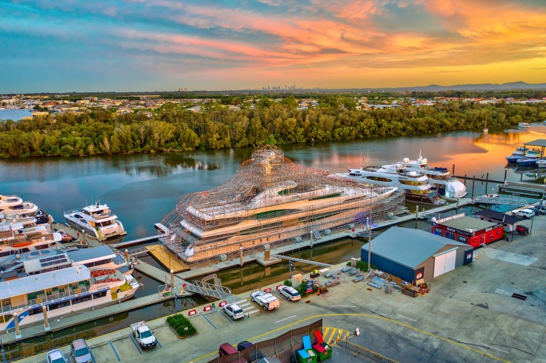 A marina at sunset with several yachts moored, including a large yacht under construction covered in scaffolding. The scene includes parked cars, buildings, trees, and a city skyline in the distance.
