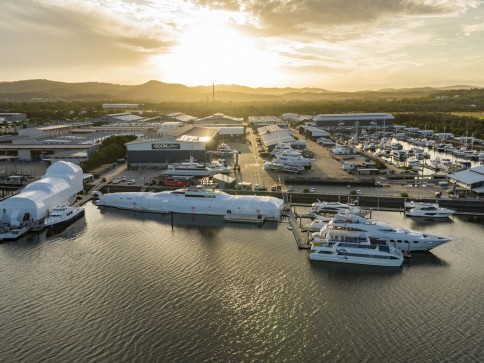 Aerial view of a marina at sunset showing several large yachts moored at piers, with industrial buildings and more boats in the background. The scene is calm with golden sunlight reflecting on the water.