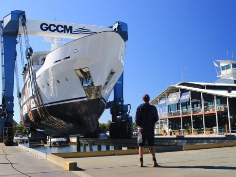A person stands on a jetty facing a large yacht being lifted out of the water by a blue GCCM boat hoist, with a marina building and clear blue sky in the background.