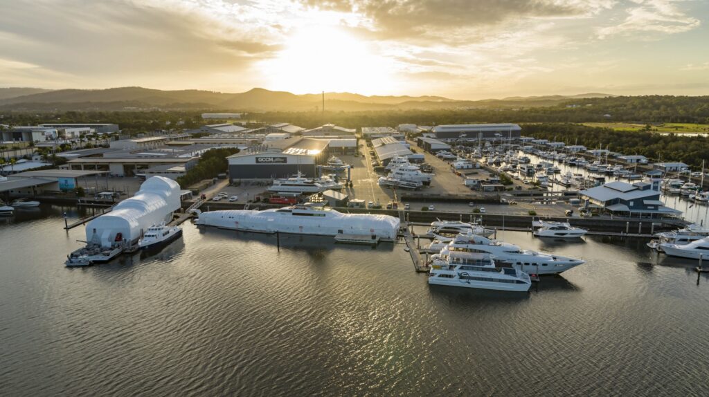 A marina at sunset with several yachts and boats moored along jetties. Large covered vessels are near the forefront, and buildings and mountains are visible in the background under a partly cloudy sky.
