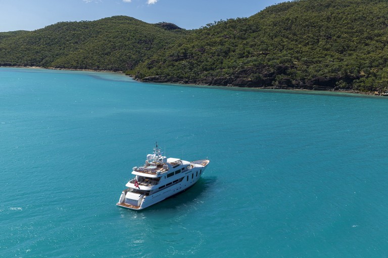A white luxury yacht floats on bright turquoise water near a lush, green, hilly coastline under a clear sky.