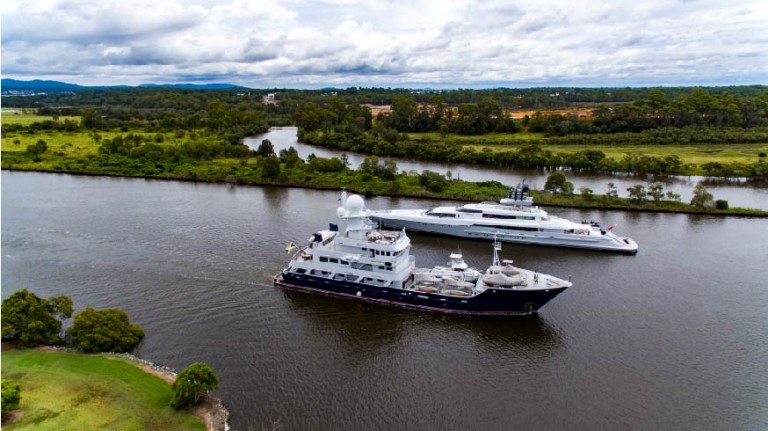 Two large yachts are sailing on a calm river with green trees, grass, and cloudy skies in the background, creating a scenic, peaceful landscape.