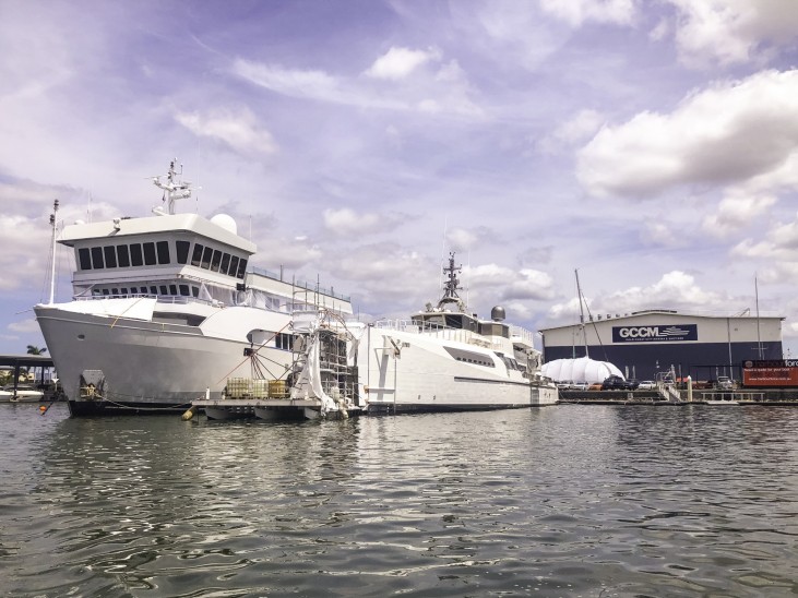 Two large white ships are moored at a marina near a building labelled GCCM under a partly cloudy sky. The water is calm, reflecting the vessels and nearby structures.