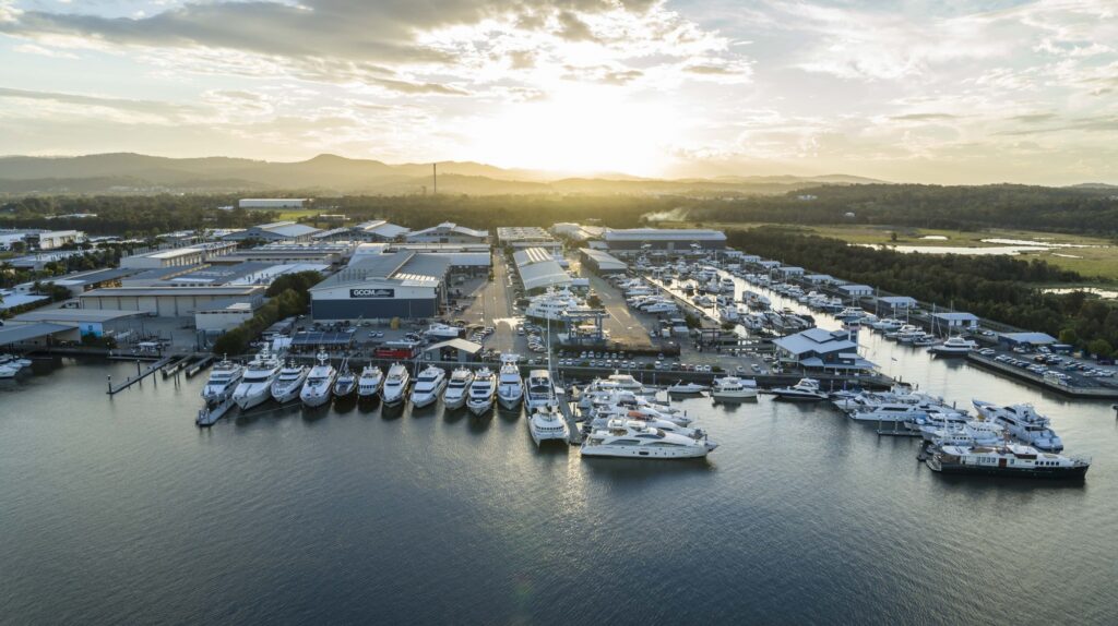 Aerial view of a marina at sunset with numerous yachts and boats moored along piers, surrounded by industrial buildings, greenery, and distant hills under a partly cloudy sky.