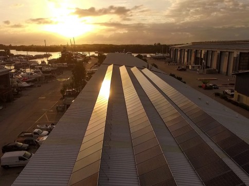 Rows of solar panels cover the roof of a large industrial building at sunset, reflecting sunlight. Nearby are parked vehicles, boats, and warehouses, with water and trees visible in the background.