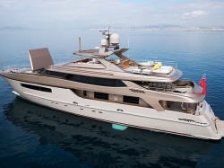A modern luxury yacht with a sleek design floats on calm blue water. The yacht features multiple decks, a shaded seating area, and a red flag at the stern. The coastline is visible in the background under a partly cloudy sky.