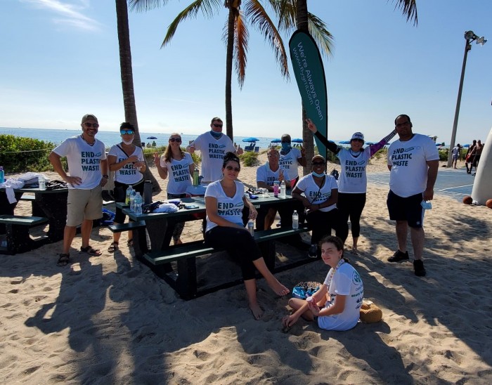 A group of people wearing “End Plastic Waste” shirts pose together on a sunny beach near picnic tables, with palm trees, the sea, and blue sky in the background. They appear to be taking part in a cleanup event.
