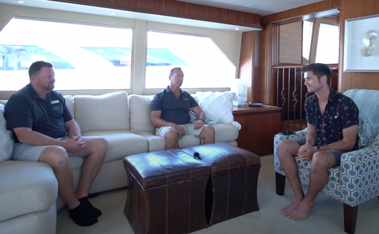 Three men are having a conversation in a well-lit, modern room with large windows on a yacht. Two are seated on a white sofa and one is sitting barefoot in an armchair. A leather footstool with a microphone sits between them.