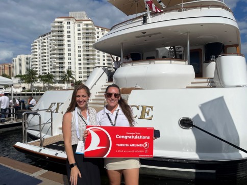 Two women stand smiling in front of a large white yacht, holding a red and white Congratulations Turkish Airlines sign. Tall buildings and palm trees are visible in the background under a partly cloudy sky.