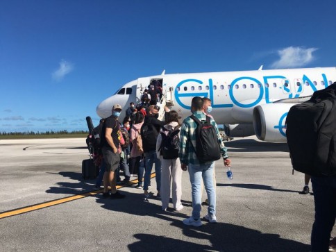 Passengers wearing rucksacks and face masks stand in a queue and board a white and blue aeroplane on a sunny day, with clear skies and some trees visible in the background.
