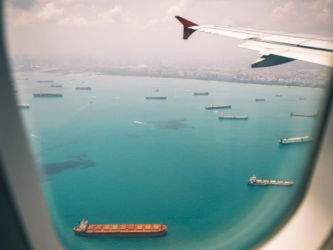 View from an aeroplane window showing several large cargo ships scattered across turquoise water near a coastline, with part of the aeroplane wing visible against a hazy sky.