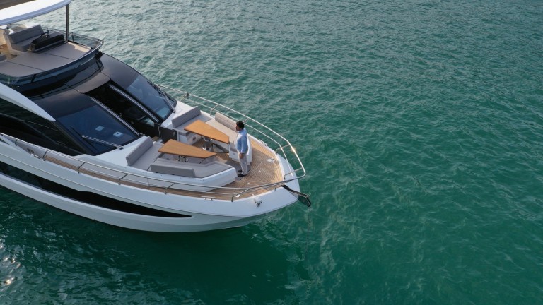 A person stands on the bow of a modern white yacht, looking out over calm, turquoise water. The yacht has outdoor seating and is partially shown, with the water surrounding it.