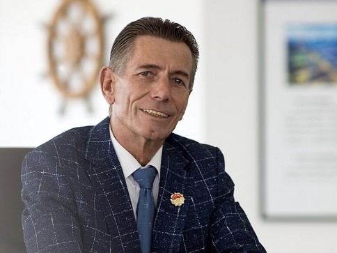A man in a blue checked suit and blue tie sits and smiles in an office setting, with a blurry framed picture and ship’s wheel visible in the background.
