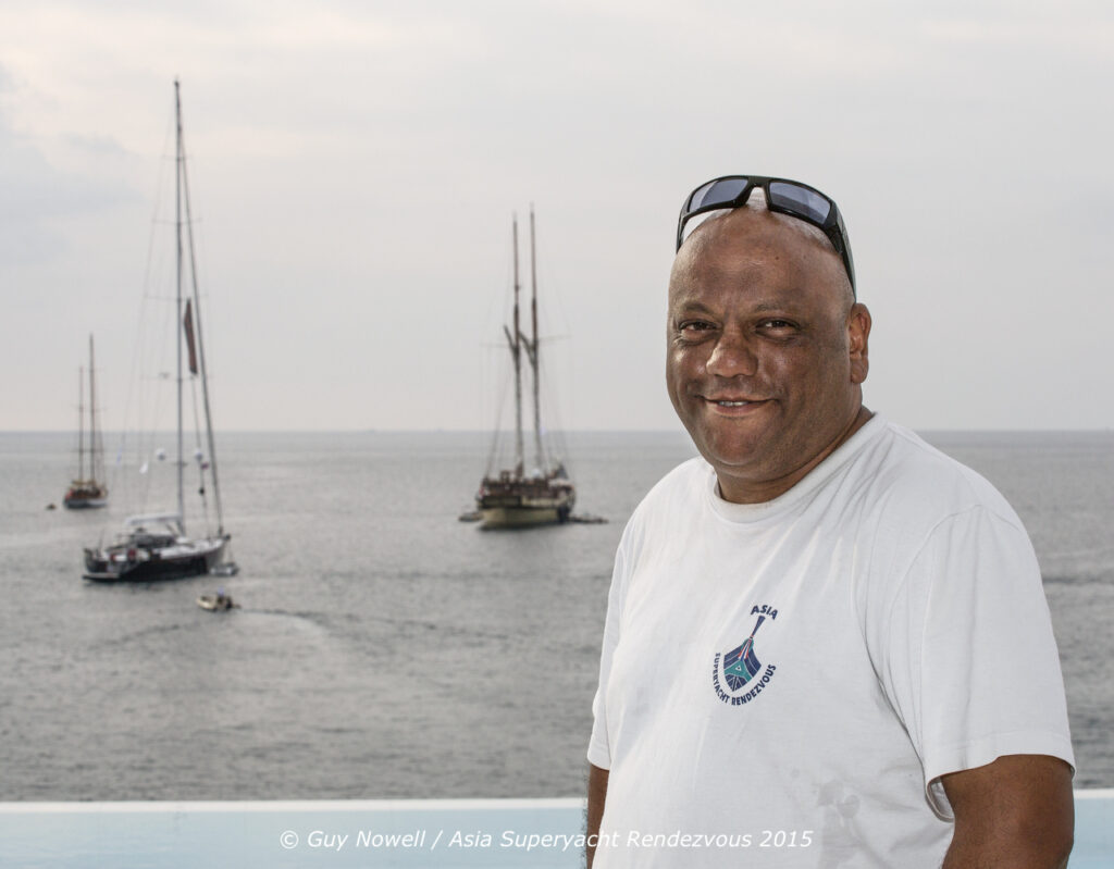A man in a white T-shirt stands smiling by the waterfront with several sailboats anchored on the calm sea behind him. The sky is overcast. Text at the bottom credits the photo to Guy Nowell, Asia Superyacht Rendezvous 2015.
