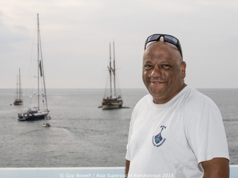 A man wearing a white T-shirt and sunglasses on his head stands smiling by the sea, with several sailing boats in the background under a cloudy sky.