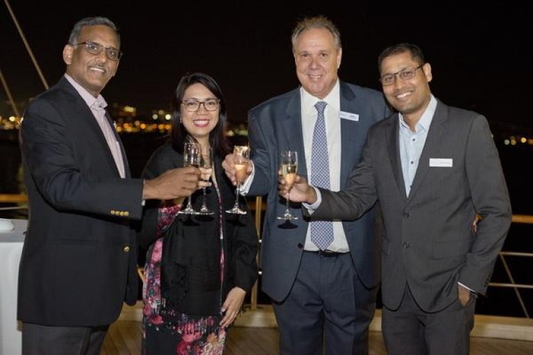 Four people in formal attire stand on a deck at night, smiling and clinking champagne glasses, with city lights visible in the background.