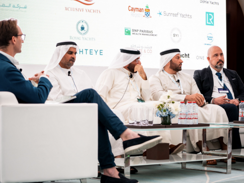 Five men sit on a stage during a panel discussion; four wear traditional Middle Eastern attire and one wears a suit. Branded banners and water bottles are visible, indicating a formal event or conference.