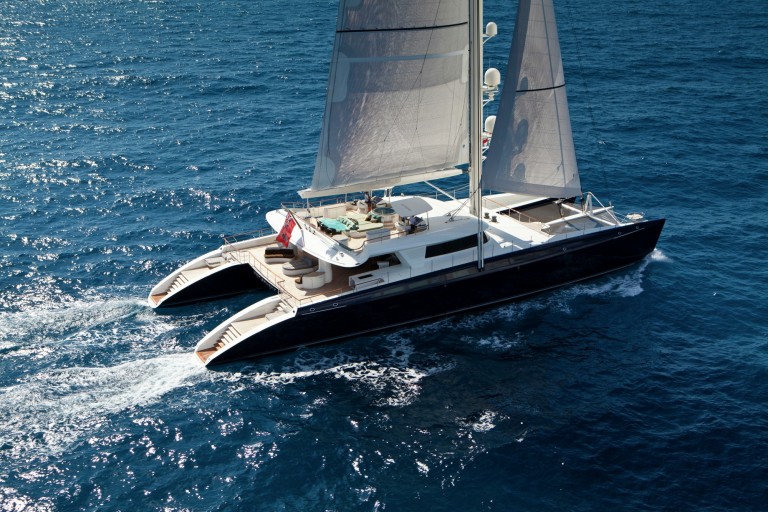 A large, modern catamaran yacht with white sails glides across deep blue ocean water, leaving a wake behind. The deck features seating areas and people can be seen relaxing on board.