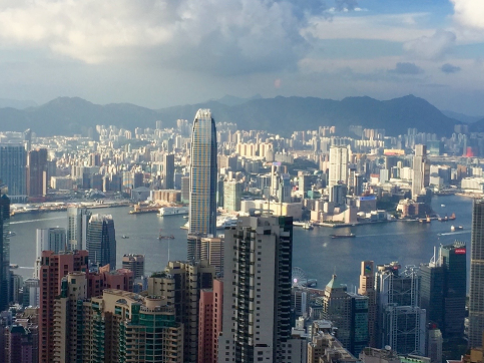 A panoramic view of a city skyline with tall skyscrapers and a harbour, set against distant mountains under a partly cloudy sky.