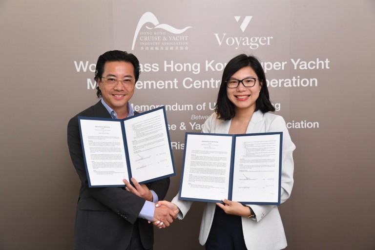 A man and a woman in business attire smile whilst shaking hands and holding signed documents in front of a backdrop with company logos and text about a memorandum of understanding.