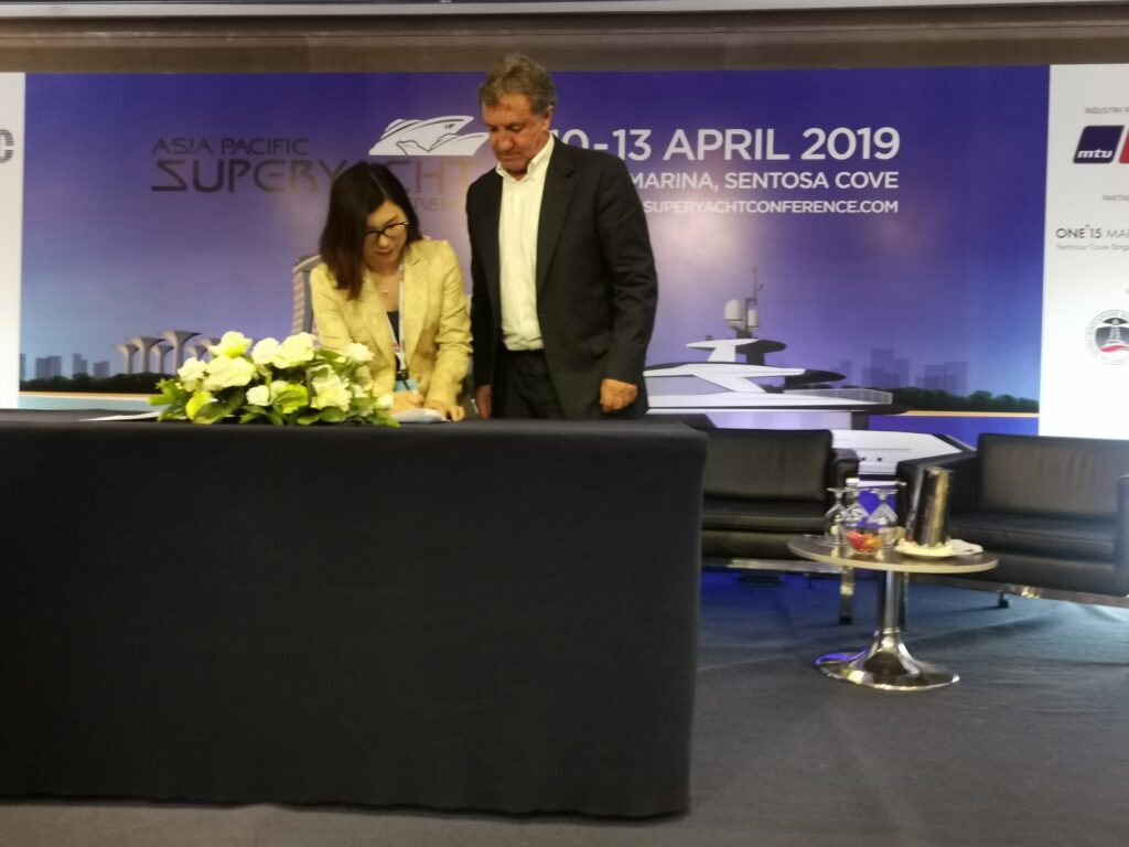 A woman signs a document at a table with flowers, as a man stands beside her. Behind them is a banner for the Asia Pacific Superyacht Conference 2019 at ONE°15 Marina, Sentosa Cove.