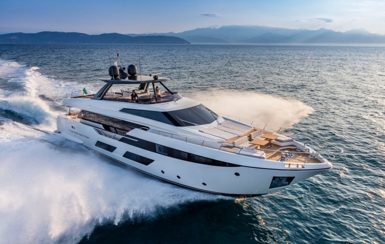A modern white luxury yacht speeds across the blue sea, creating waves as it moves. The yacht has sleek lines, an open deck with seating, and mountainous land visible in the background under a clear sky.