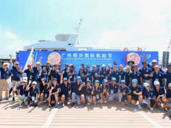 A large group of people dressed in navy shirts and caps pose together on a dock in front of a boat, cheering and smiling under a sunny sky with a blue event banner in the background.