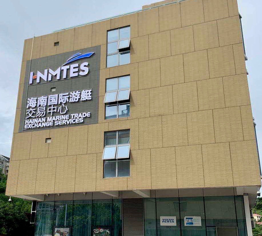 A beige building with large signs reading INMTES and HAINAN MARINE TRADE EXCHANGE SERVICES in English and Chinese. Several windows are open, and the ground floor has glass walls.