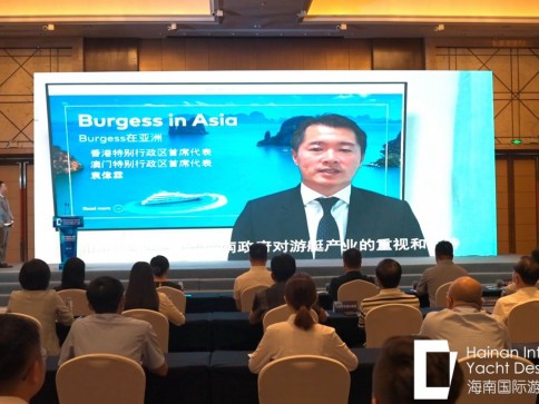 A man in a suit speaks on a large screen during a conference. Attendees sit facing the screen in a formal setting, with a presentation slide behind the speaker titled Burgess in Asia.