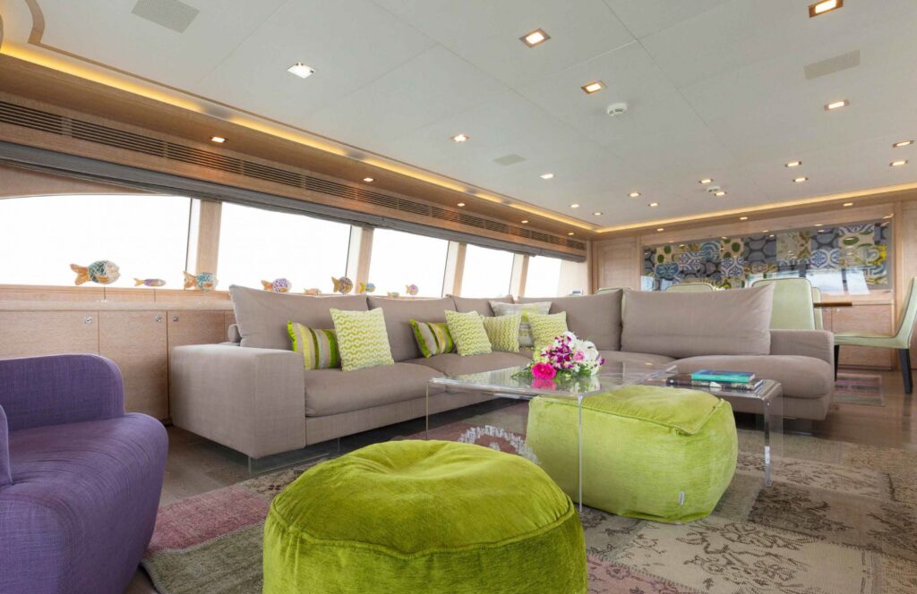 A modern yacht lounge with large windows, beige sectional sofa, green pouffes, purple armchair, glass coffee table with flowers, and decorative cushions in shades of green and yellow. Artwork and fish sculptures decorate the space.