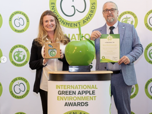 Two people stand smiling at an awards event. The woman holds a trophy, and the man holds a certificate. A large green apple sculpture is on the table between them, with award banners in the background.
