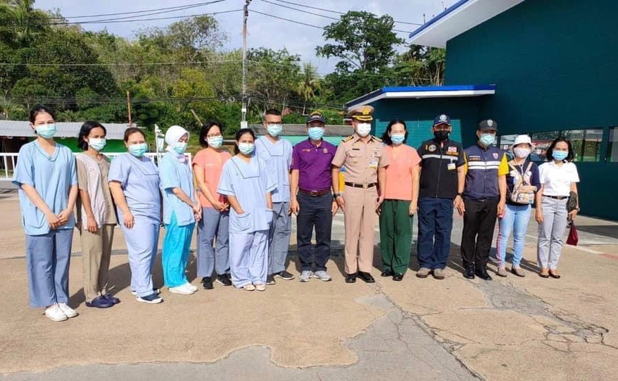 A group of people, including medical staff in scrubs, police officers, and others, stand outdoors in a queue, all wearing face masks. Trees and a building are visible in the background.