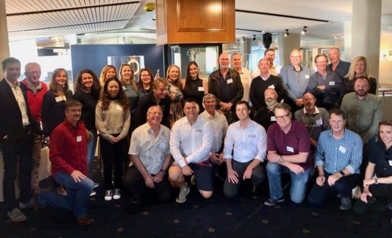 A group of about 30 adults, both men and women, pose together indoors for a photo. Most are standing, with a few kneeling in front. They are smiling and wearing name badges, suggesting a professional or social event.
