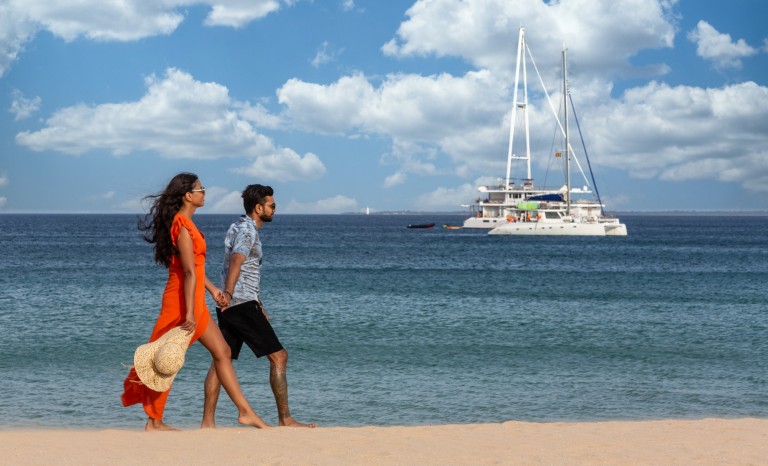 A couple walk hand-in-hand along a sandy beach, with the woman in an orange dress holding a sunhat. The sea is calm and a white catamaran floats in the background under a blue sky with scattered clouds.