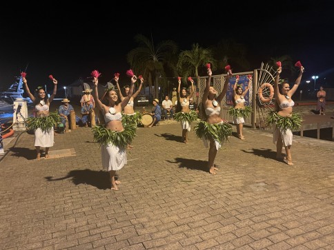 A group of women in traditional Polynesian attire perform a dance outdoors at night, holding red pom-poms, with musicians playing drums in the background under palm trees.