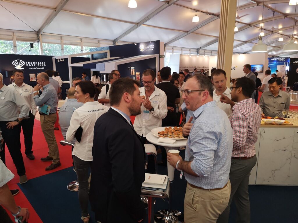 A busy indoor networking event or conference with people in business attire talking in groups, standing around high tables with snacks, under a large marquee with company stands and banners in the background.