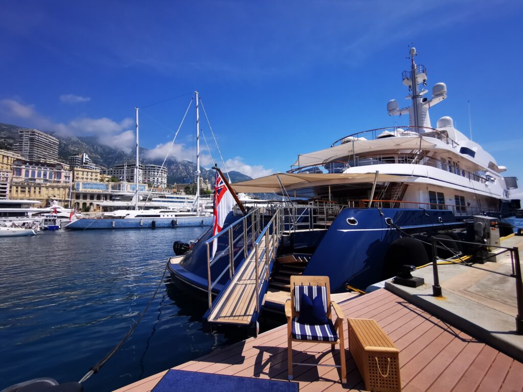 A large luxury yacht moored at a marina on a sunny day, with a gangway leading to the deck, nearby lounge chairs and tables, other boats berthed in the background, and buildings along the waterfront.