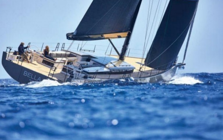 A modern sailboat named BELLA sails on bright blue ocean waves under a clear sky, with two people relaxing on deck. The boat has sleek black sails and a wooden deck.
