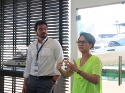 Two people stand indoors near large windows with boats visible outside. One person in a white shirt listens whilst the other, wearing glasses and a bright yellow-green shirt, gestures whilst speaking.