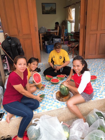 Four women sit on a tiled floor, smiling and preparing watermelon slices. Watermelons and bags are scattered around them. A man is seen in the background inside the room. The atmosphere appears casual and friendly.