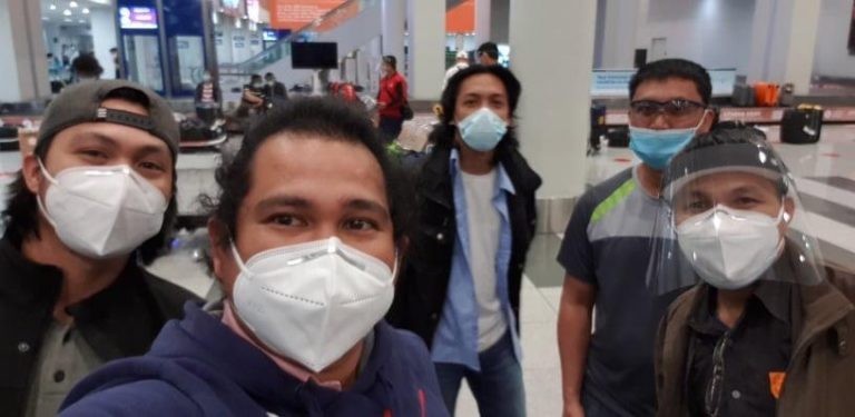 Five men wearing face masks and some with face visors pose for a selfie inside an airport baggage reclaim area, with luggage and other passengers visible in the background.