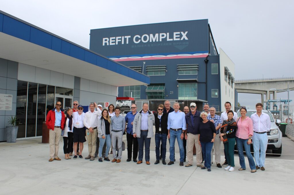 A group of around twenty people pose together outside a large blue building labelled Refit Complex on a cloudy day. Some are smiling, and the group appears to be diverse in age and attire.