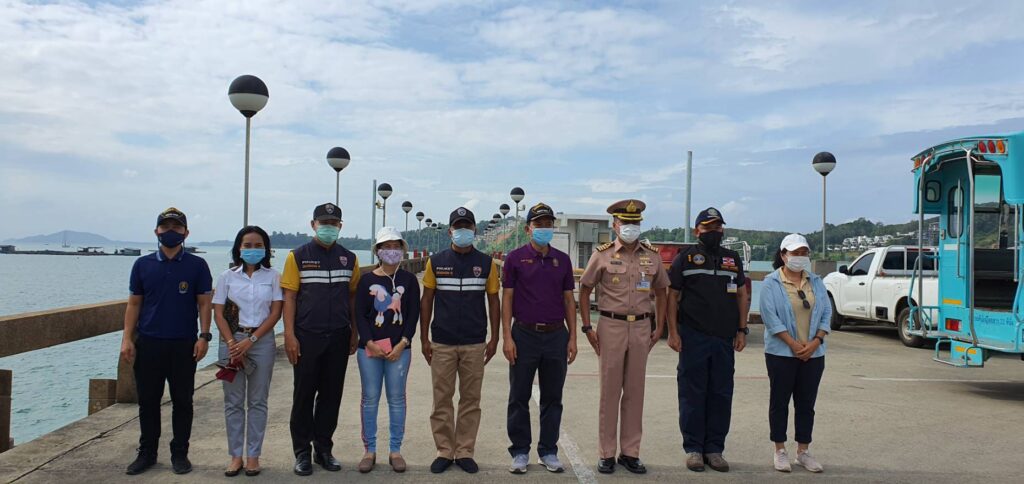 A group of nine people, including men and women, stand in a row on a pier near the sea. Most are wearing uniforms, all are wearing masks, and a blue vehicle is parked nearby under a partly cloudy sky.
