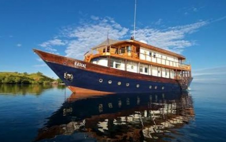 A large wooden yacht named RASCAI floats on calm, reflective water under a blue sky with light clouds. The boat has multiple decks and is surrounded by scenic greenery in the background.