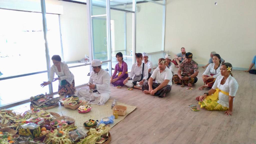 A group of people in traditional Balinese attire sit on the floor, participating in a ceremonial ritual with offerings arranged on woven mats in a bright room with large windows.